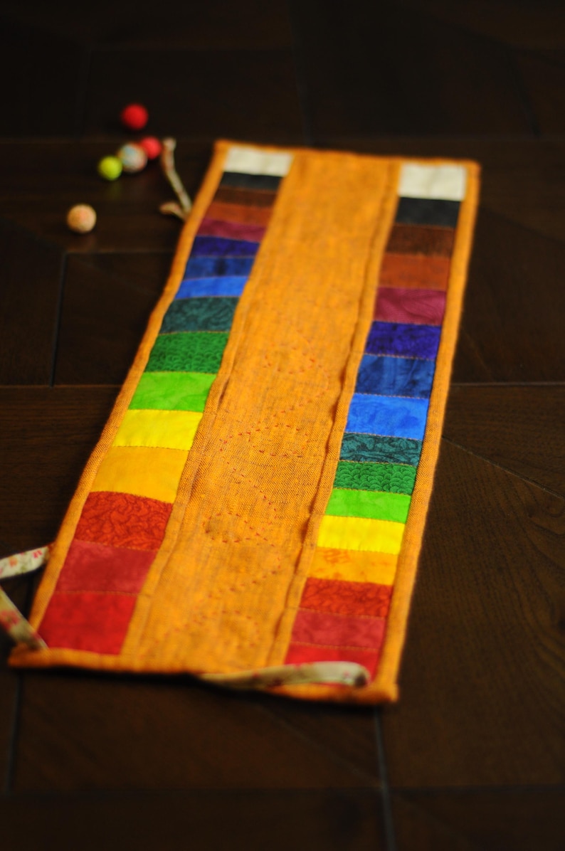 May include: A long, orange fabric pouch with a rainbow of colors sewn along the edge. The pouch is folded in half and has a white string tie.
