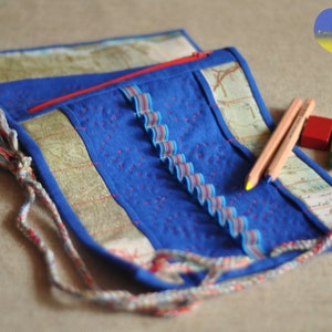 May include: A blue fabric pencil roll with a zipper closure and a colourful tassel. The roll is made of different fabrics and has a decorative ribbon sewn along the edge. Two wooden pencils and a few coloured blocks are visible in the image.