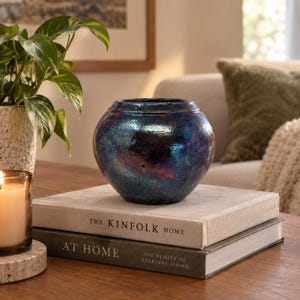 May include: A round, iridescent ceramic vase with a deep blue and purple metallic glaze, featuring a textured, speckled finish. The vase sits atop two stacked hardcover books titled The Kinfolk Home and At Home, The Beauty of Everyday Living.