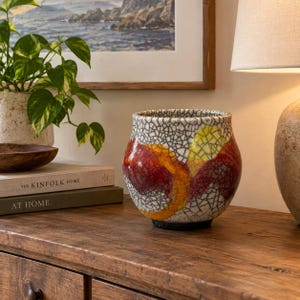 May include: A ceramic planter with a white base featuring a black crackle glaze pattern and bold, abstract swirls of red, orange, and yellow. The vessel has a rounded, bulbous shape with a wide opening and a dark, solid-colored circular base.