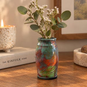 May include: A small ceramic vase with a glossy, multi-colored glaze featuring swirling patterns of teal, green, and burnt orange. The vase has a cylindrical body with a slightly flared rim and contains a sprig of green eucalyptus and small white flowers.