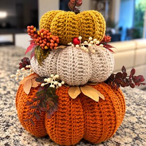 May include: A stacked set of three crocheted pumpkins in mustard yellow, beige, and burnt orange. The pumpkins are decorated with artificial orange berries, white berries, and dark red leaves tucked between the layers of yarn.