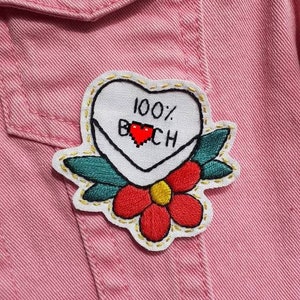 May include: A pink denim jacket with a white embroidered patch that says "100% B*CH" with a red heart. The patch is surrounded by green leaves and a red flower with a yellow centre.
