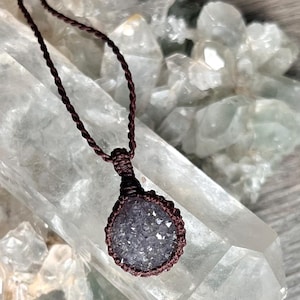 May include: A dark purple druzy gemstone pendant secured in a brown macrame setting. The pendant hangs from a matching brown braided cord, featuring a circular stone with a textured, crystalline surface that reflects light across its facets.