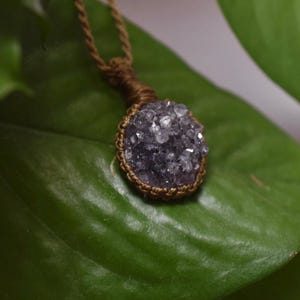 May include: A pendant necklace featuring a circular, deep purple druzy crystal stone encased in a brown macrame cord setting. The crystal has a textured, sparkling surface, and the pendant hangs from a matching brown twisted cord necklace.