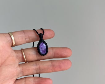 Könnte beinhalten: Eine Halskette mit einem ovalen, violetten Steinanhänger, der in einer schwarzen Makramee-Fassung befestigt ist. Der Anhänger hängt an einer dünnen schwarzen Kordel und zeigt einen tiefvioletten Farbton mit natürlichen weißen Adern.