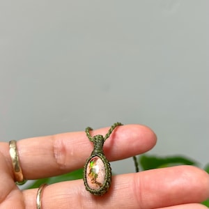 May include: An oval-shaped pendant featuring a speckled beige stone with iridescent green and orange flecks, encased in a dark olive green macrame cord setting. The pendant is attached to a matching thin olive green cord necklace.