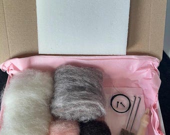 Op de afbeelding: Een naaldvilten set met vier bollen wol in wit, grijs, roze en bruin, twee vilten naalden, een klein borsteltje en een vilten gereedschap. De set is verpakt in een roze doos met wit tissuepapier.