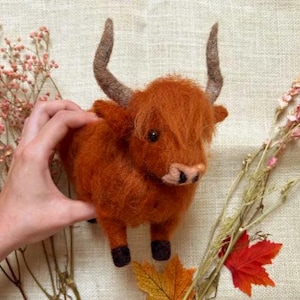 May include: A handcrafted, orange-brown felt Highland cow figurine with small, curved horns and black hooves. The cow is surrounded by dried flowers and fall foliage, creating a rustic, autumnal theme.