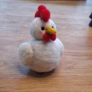 May include: A handcrafted white felt chicken figurine with a red comb and wattle, and a yellow beak. The chicken has small black eyes and rosy cheeks, and is sitting on a wooden surface.