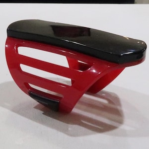 Puede incluir: Una protección de mano de plástico rojo con una parte superior negra. La protección tiene una serie de ranuras horizontales en el lateral.