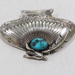 Puede incluir: Un broche o colgante decorativo plateado y artesanal. El diseño presenta una piedra de turquesa ovalada central, rodeada por un patrón en forma de abanico, plumas y una cornucopia. La pieza tiene una estética antigua o vintage.