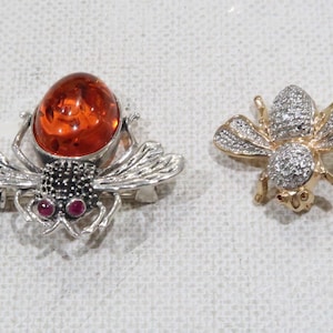 Puede incluir: Dos broches de abeja, uno con una piedra de ámbar y detalles rojos, el otro con piedras transparentes.