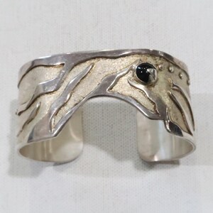 Könnte beinhalten: Silbernes Armreif-Armband mit strukturiertem Design und einem schwarzen Edelstein. Das Armband hat ein breites Band mit offenem Design. Das Metall hat ein gebürstetes Finish mit einem kontrastierenden strukturierten Zentrum.