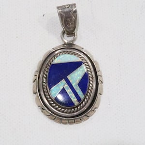 Pode incluir: Pingente oval de prata com uma pedra central azul escuro incrustada com formas geométricas brancas e azul claro. O pingente tem uma borda decorativa prateada e uma argola prateada.