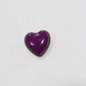 May include: A heart-shaped brooch with a glossy, deep purple center. The brooch is framed by a silver-colored, twisted rope-like border. The jewelry piece is set against a white background.