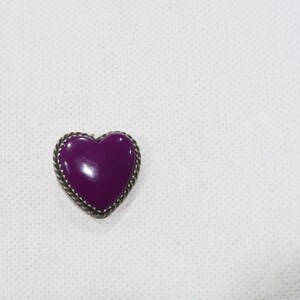 May include: A purple heart-shaped brooch with a silver-colored rope-like border. The heart is a deep, glossy purple. The brooch is set against a white, textured background.