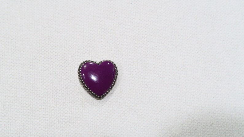 May include: A purple heart-shaped brooch with a silver-colored rope-like border. The heart is a deep, glossy purple. The brooch is set against a white, textured background.