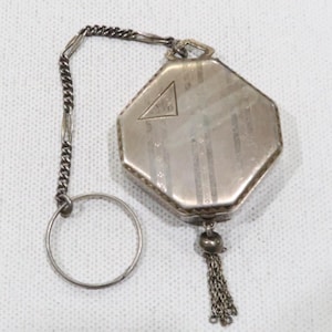 Puede incluir: Un reloj de bolsillo octogonal plateado con cadena y anillo. La esfera del reloj tiene un emblema triangular y un patrón de rayas verticales. Una pequeña borla cuelga de la parte inferior. La cadena está conectada a un anillo.