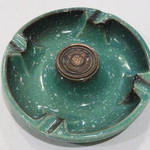 Puede incluir: Un cenicero de cerámica verde con un esmalte blanco moteado y una pieza central de color cobre. La pieza central tiene un diseño en relieve con las palabras "The American Legion" alrededor del borde.