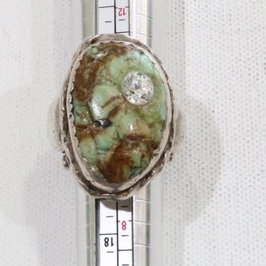 May include: Oval-shaped silver ring featuring a turquoise stone with brown veining and a central clear, round gemstone. The ring is set on a measuring tool, showing size markings.