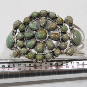 Könnte beinhalten: Ein silbernes Armreif-Armband mit einem zentralen kreisförmigen Design, das mit grünen und hellbraunen Steinen verziert ist. Das Armband hat zusätzliche Steine an den Seiten und aufwendige Metallarbeiten. Ein Schmuckstück.