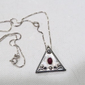 May include: A silver necklace with a triangular pendant featuring a red gemstone and intricate silver detailing.