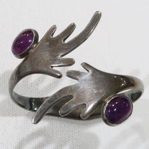 May include: A silver bracelet with two purple gemstones set into the design. The bracelet is shaped like two hands reaching out to each other.