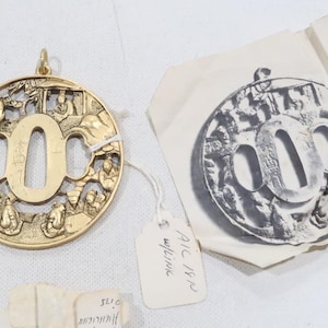 May include: Gold-colored metal pendant with a large, open 'O' in the center. The pendant is round and features a detailed design of figures and symbols around the 'O'.