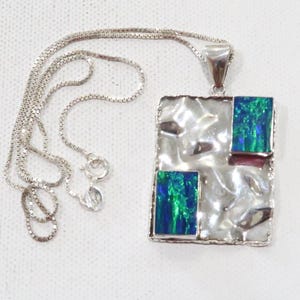 Puede incluir: Un collar de plata con un colgante rectangular. El colgante tiene una superficie plateada texturizada y martillada con dos incrustaciones rectangulares de material iridiscente azul y verde. La cadena del collar es una fina cadena de caja.