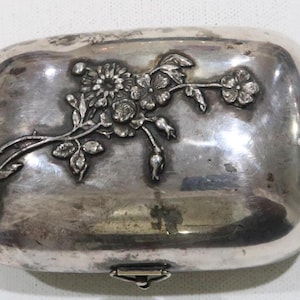 May include: A silver-toned, rectangular trinket box with a floral design embossed on the lid. The box has a hinged lid and a small clasp.