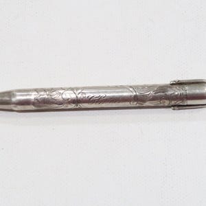 Könnte beinhalten: Ein silberner Bleistift mit aufwendigen floralen Gravuren. Der Bleistift hat eine spitze Spitze und eine kleine Schlaufe am Ende. Der Bleistift ist aus Metall und hat ein glänzendes Finish. Es ist ein Vintage-Schreibwerkzeug.