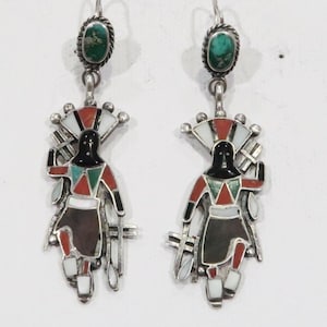 May include: A pair of silver earrings featuring a turquoise oval stone at the top. The earrings have a detailed design of a figure with a black head, red and green accents, and a fan-shaped headdress. The figure is wearing a skirt with brown, white, and red details.