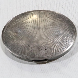 May include: A silver oval compact with a sunburst design and the initials "A.E.S." engraved on the front.