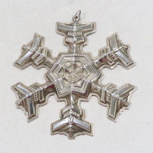 May include: A silver snowflake Christmas ornament with a detailed design. The snowflake has six points and a loop at the top for hanging.