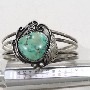 May include: A silver bracelet featuring a turquoise stone set within an ornate silver design. The bracelet is crafted from multiple bands of silver and incorporates a leaf-like motif on one side.