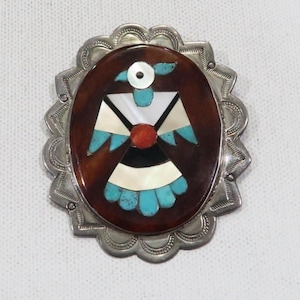 May include: Oval silver-tone brooch featuring a bird design. The bird is crafted from turquoise, white, black, and red stones, set against a brown backdrop. The brooch has a scalloped edge.