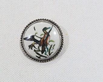 May include: A silver-toned brooch with a white porcelain cameo featuring a hand-painted image of a duck in flight.