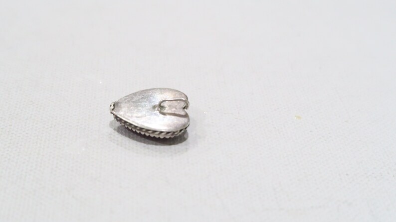 May include: A small, silver-colored heart-shaped pendant. The pendant has a textured surface and a decorative rope-like border. A small loop is attached to the top for hanging. The pendant is likely made of metal and is suitable for jewelry making.