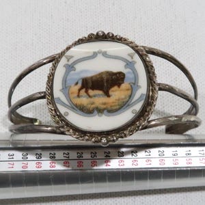 Op de afbeelding: Zilverkleurige manchetarmband met een centraal rond medaillon met een afbeelding van een bizon in een veld. Het medaillon is omlijst door een decoratieve rand en geplaatst tegen een witte achtergrond. Het ontwerp van de armband omvat meerdere gebogen banden.