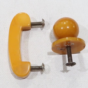 May include: Two vintage amber bakelite cabinet knobs. One is a round knob with a flat base and the other is a curved handle.