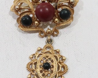 May include: A gold-toned brooch featuring a red and black stone design. The brooch has a dangling pendant with a black stone at its centre.