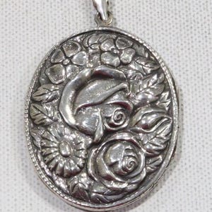 May include: Oval silver pendant with a floral design. The pendant features a raised relief of a rose, other flowers, and leaves. The bail is at the top for attaching to a chain. The pendant is likely made of sterling silver.