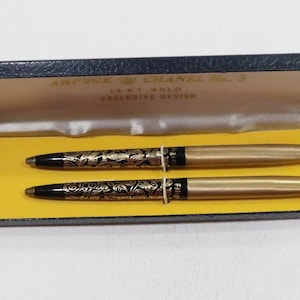 May include: A vintage pen set in a blue case with gold trim. The case is open, revealing two pens with black and gold floral designs. The pens have gold-colored barrels and are displayed on a yellow base.