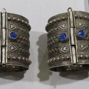 Può includere: Un paio di braccialetti in argento con cerniera, con disegni intricati e pietre preziose blu. I braccialetti sono decorati con piccole perline e cuori.