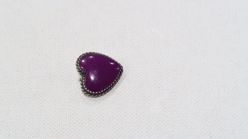 May include: A heart-shaped purple brooch with a silver-colored rope-like border. The brooch is set against a white background. The heart is a solid, glossy purple color.