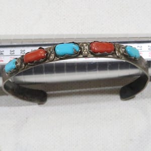 Puede incluir: Un brazalete de plata con piedras turquesa y coral. El brazalete está hecho de una fina banda de plata con un diseño decorativo. Las piedras están engastadas en engastes de plata y están dispuestas en un patrón repetitivo.