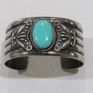 May include: A silver cuff bracelet with a turquoise stone set in the center. The bracelet has a decorative design with a repeating pattern of small circles and a floral motif.