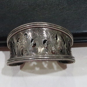 May include: A silver bracelet with an intricate floral design. The bracelet is wide and has a rounded shape.
