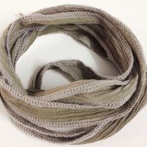 Könnte beinhalten: Ein aufgerolltes olivgrünes Band mit knittriger Textur, das an beiden Seiten mit einer hellgrauen Kettelnaht versehen ist. Das Band ist zu einer kreisförmigen Schleife gelegt und zeigt den Kontrast zwischen dem Stoff und der Naht.